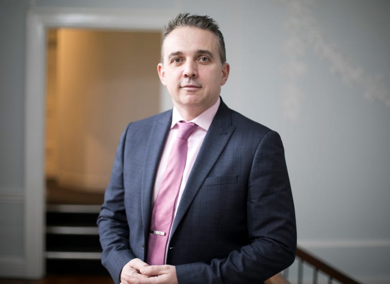 Dermot Wells, wearing a navy suit and pink tie, standing in a formal indoor setting.