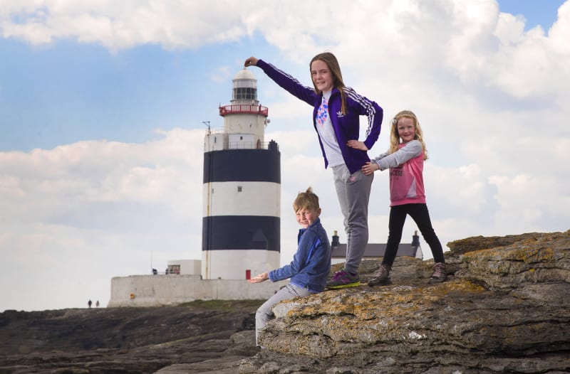 Three children posing on rocks in front of Hook Lighthouse, a black and white striped historic lighthouse in Ireland.