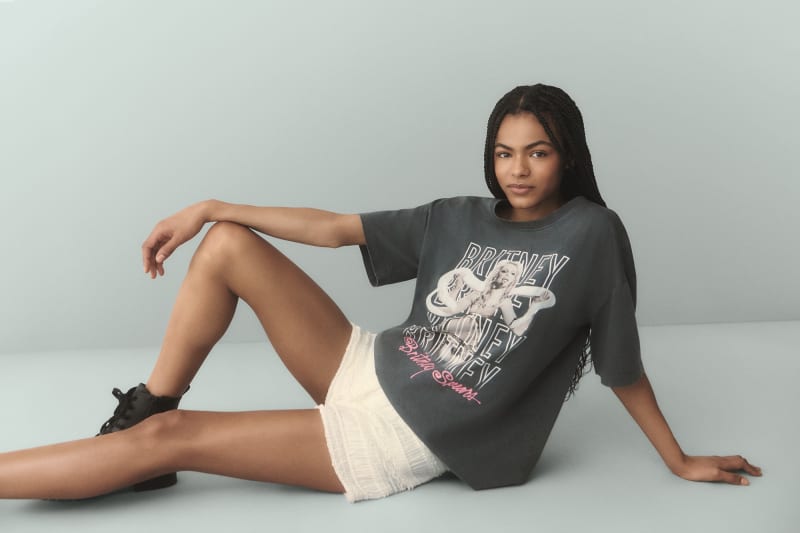 Model wearing oversized dark grey Britney Spears graphic t-shirt with white text and pink script logo, white shorts, black sneakers.