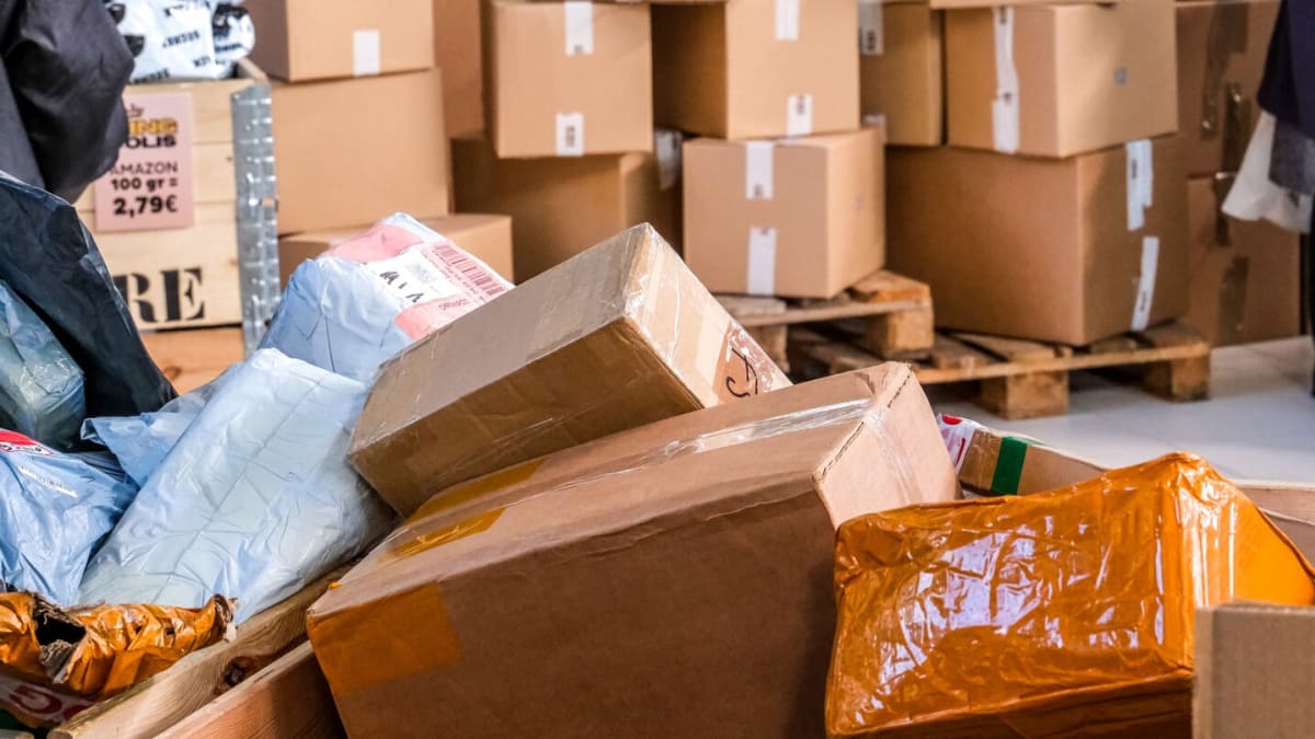 Pile of mixed parcels and damaged packages in a warehouse with stacked cardboard boxes on pallets behind.