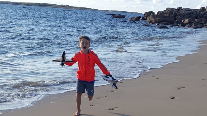 Samuel Roquette running barefoot on a beach holding a toy airplane and shoes, laughing with excitement.