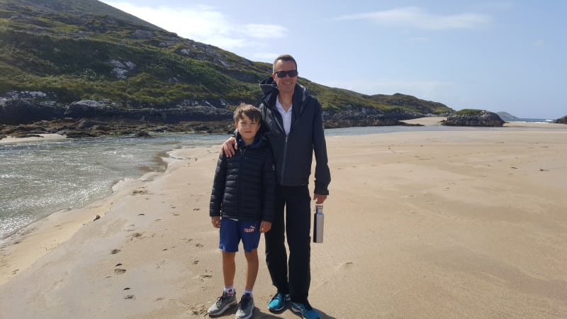 Samuel and Benjamin standing together on a sandy beach with rocky hills and a tidal stream in the background.