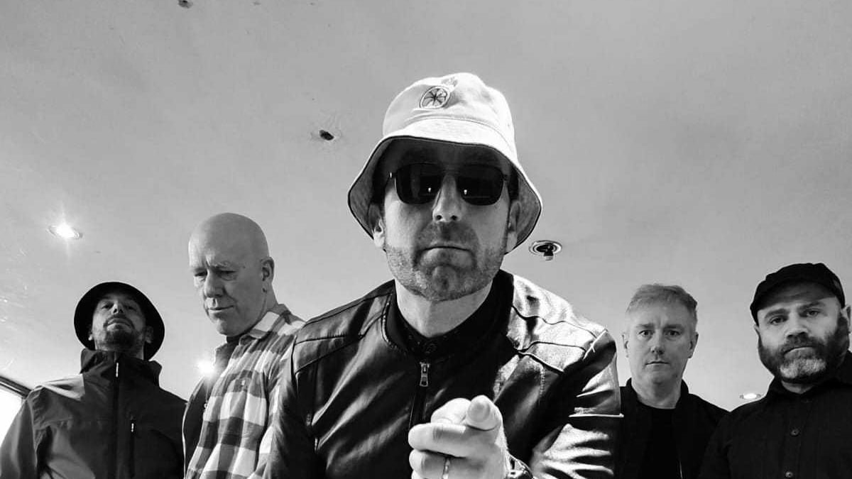 Oas1s band posed together in black and white, central member in bucket hat and sunglasses pointing at camera.