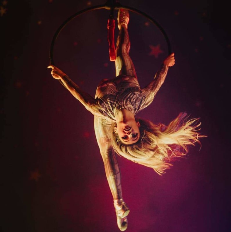 Kourtney performing an aerial hoop act, hanging upside down in a patterned bodysuit on a lit stage.