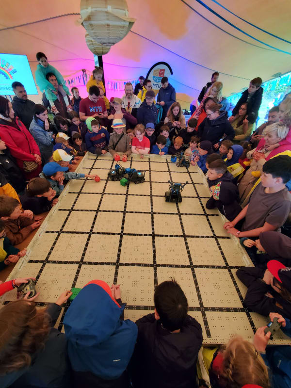 Children and adults gather around a large robot battle grid at an MTU robotics event inside a colourful tent.