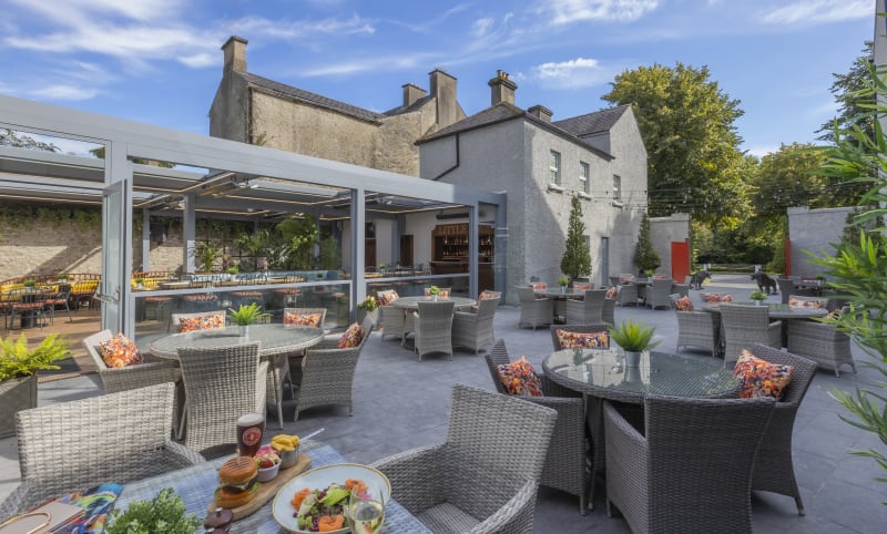 Outdoor terrace dining area with grey rattan furniture, colourful cushions, pergola bar, and Georgian stone building backdrop.