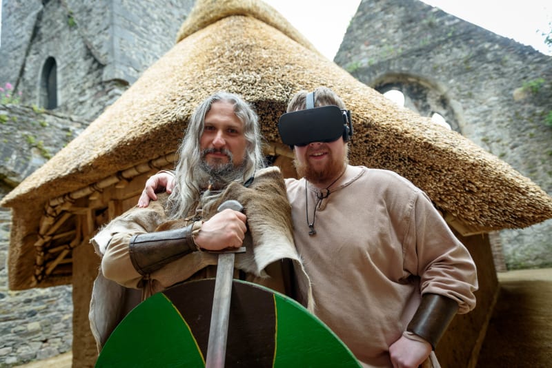 Two men in Viking-era costumes at a thatched roundhouse, one wearing a VR headset, at a castle ruin site.