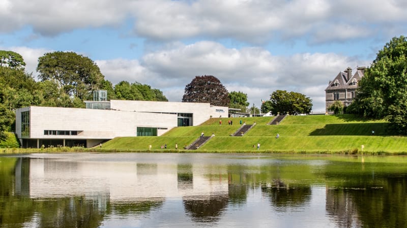 Visitors on terraces at Turlough Park museum beside a reflective lake on a cloudy summer day.