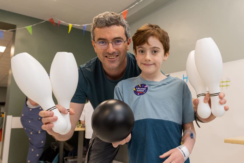 Jack and his Dad holding white bowling pins and a black bowling ball in a hospital play room, smiling at the camera.