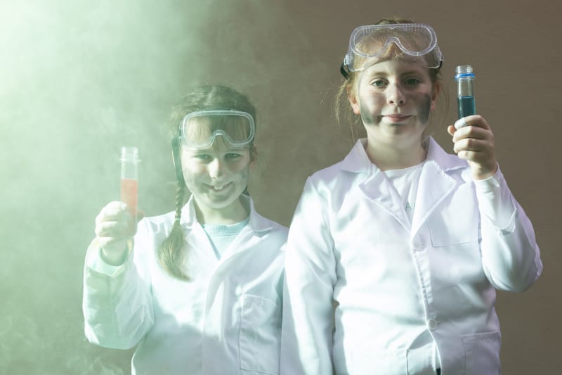 Two children in white lab coats and safety goggles hold colourful test tubes surrounded by green smoke.