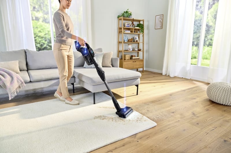 Woman vacuuming a white rug with a cordless stick vacuum cleaner in a bright modern living room.
