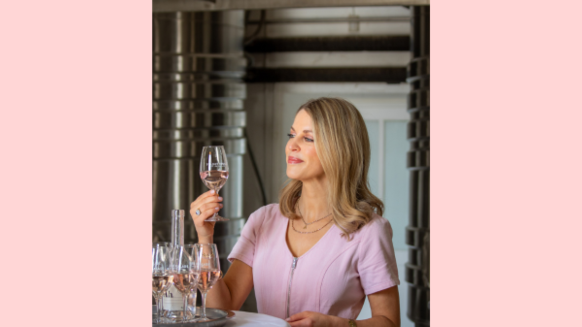 Amy examining a glass of rosé wine at a tasting table inside a winery with fermentation tanks behind her.