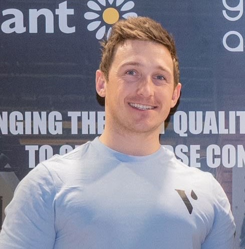 Nick Hird smiling at an event, wearing a light blue fitted t-shirt, standing in front of a branded banner.