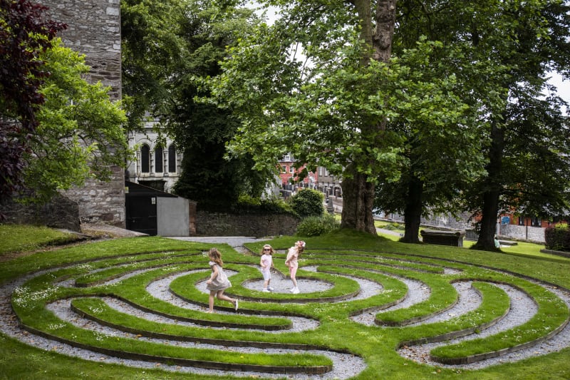 Three young girls playing in a circular grass labyrinth in a park beside a stone church in Ireland.