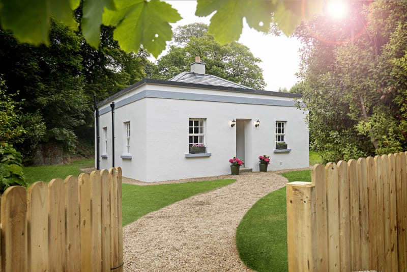 Mount Congreve Gate Lodge, a white single-storey cottage with slate roof, gravel path, wooden gate and lush garden surroundings.