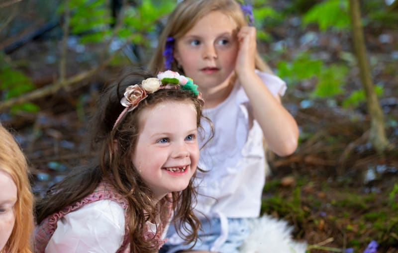 Two young girls wearing flower headbands smile and sit together in a woodland outdoor setting.