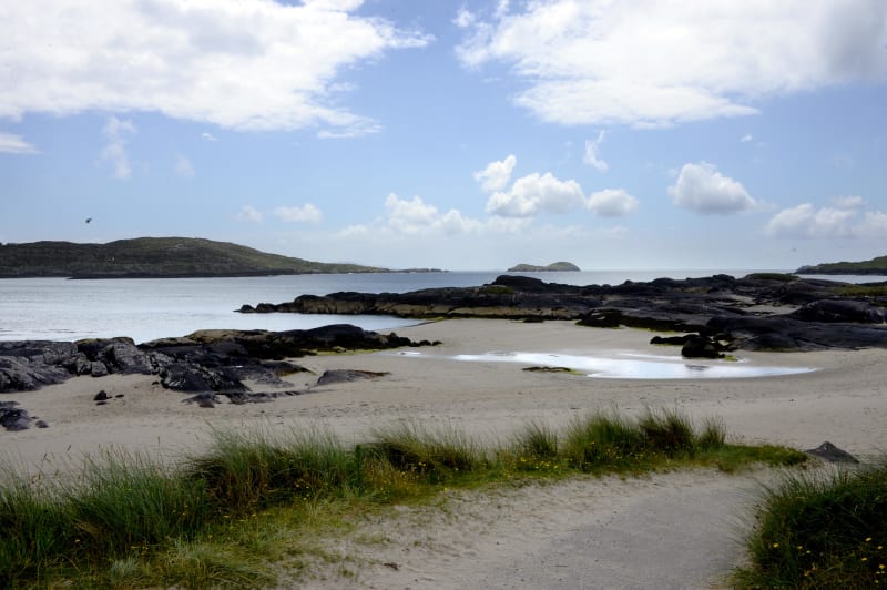 Sandy beach with dark rocks and marram grass at Derrynane, County Kerry, Ireland, with islands visible offshore.