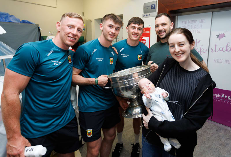 GAA players in Mayo jerseys hold Sam Maguire trophy beside a mother and baby at Temple Street Hospital.