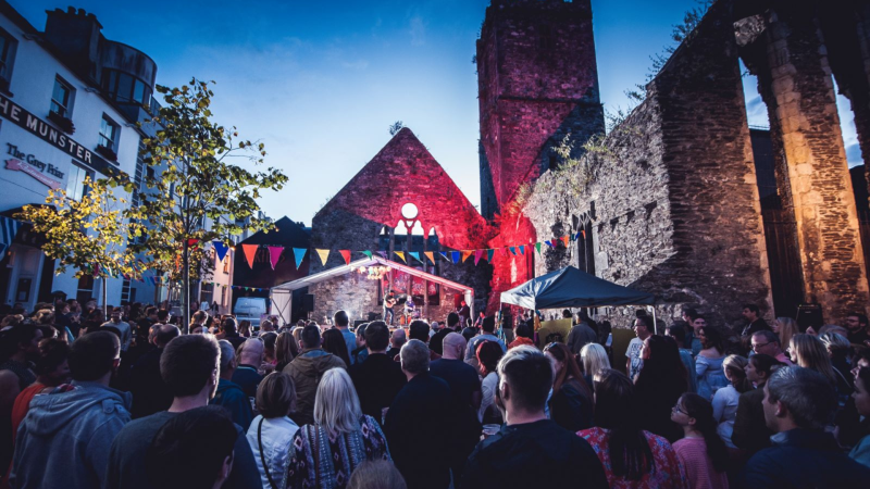 Outdoor evening music festival in a medieval churchyard with a crowd, colourful bunting and a lit stone ruin.