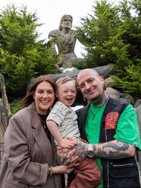 Family of three smiling in front of a large stone warrior statue at Emerald Park theme park.