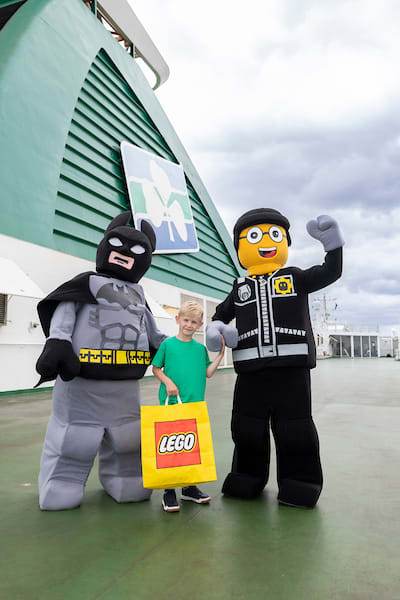 Child poses with LEGO Batman and LEGO City police officer mascots on Irish Ferries deck, holding a yellow LEGO bag.