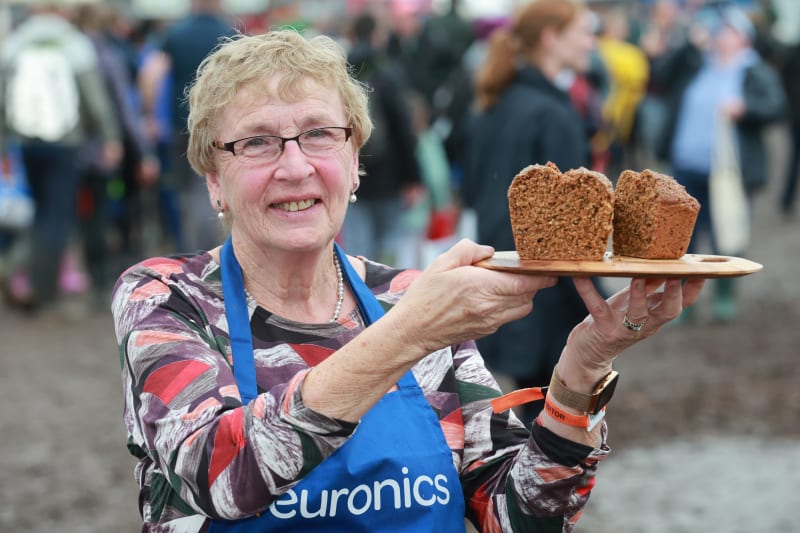Woman in Euronics apron proudly holds a wooden board with two small brown bread loaves at an outdoor event.