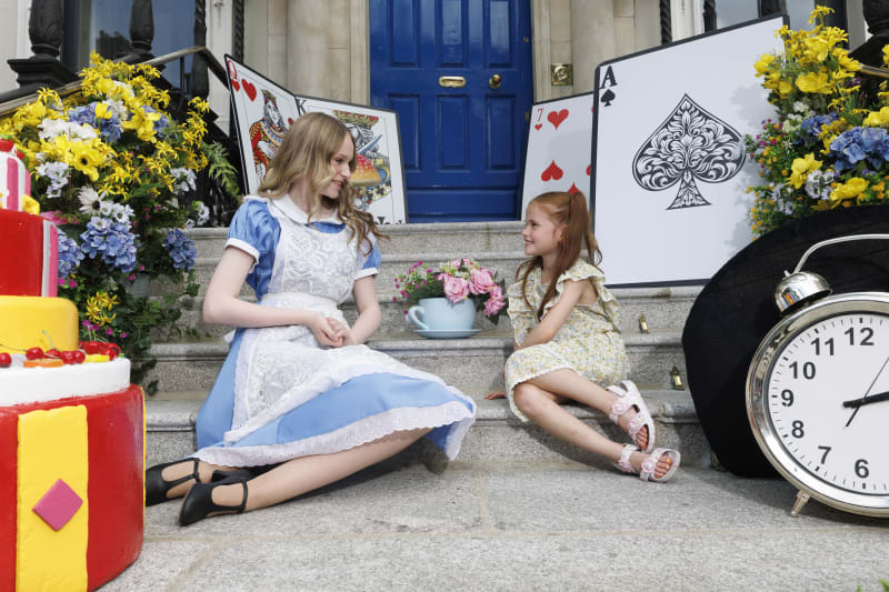 Teen dressed as Alice in Wonderland sits on steps with a young girl, surrounded by oversized Alice in Wonderland themed props.