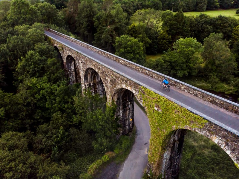 Cyclist in blue jersey rides across a historic stone viaduct surrounded by green woodland, aerial view.
