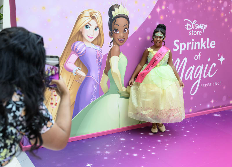 Young girl in Princess Tiana costume poses at Disney Store Sprinkle of Magic Experience backdrop with Rapunzel and Tiana illustrations.