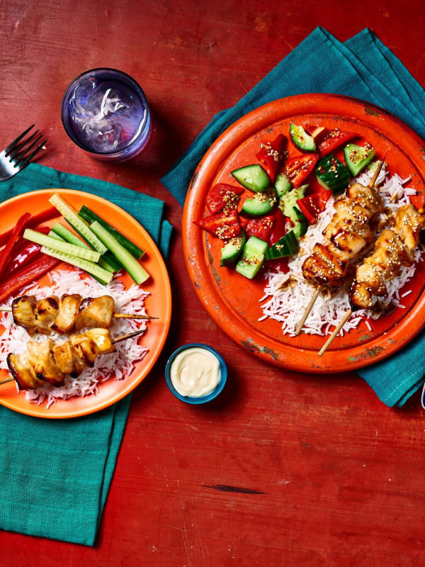 Honey sesame chicken skewers on rice with cucumber tomato salad and vegetable crudités on orange plates.