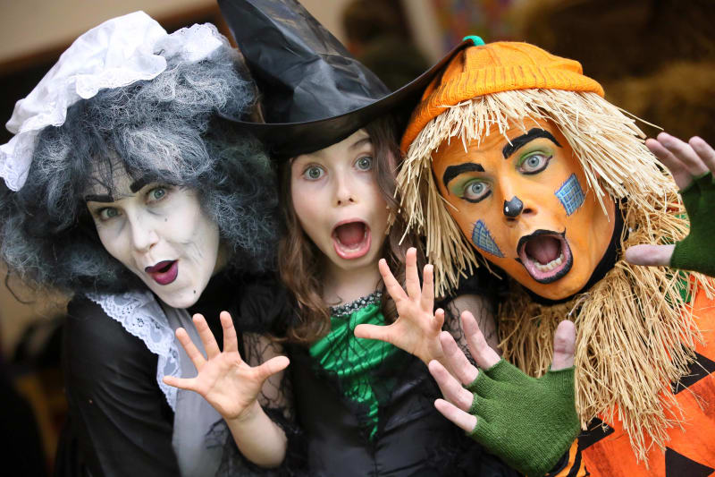 Three people in Halloween costumes — a witch, a young girl witch, and a scarecrow — pose with scary expressions.