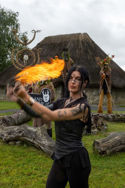 Fire performer in black with face paint spins flames at a Samhain event outside a thatched roundhouse.