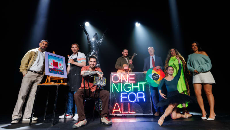 Group of performers and artists posed on stage around a neon 'One Night For All' Culture Night sign.