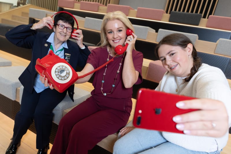 Three women pose with red communication devices — retro phone, headphones and smartphone — in a modern auditorium.