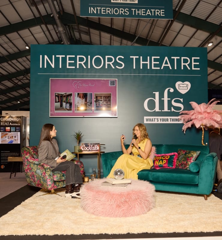 Two women at the DFS Interiors Theatre stand at a home and interiors show, one being interviewed on a teal velvet sofa.