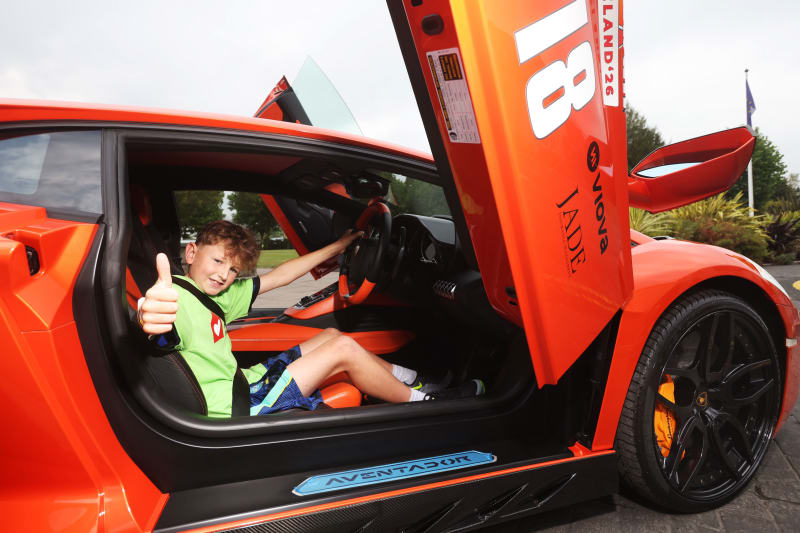 Boy sits smiling in orange Lamborghini Aventador with scissor door open, giving thumbs up.