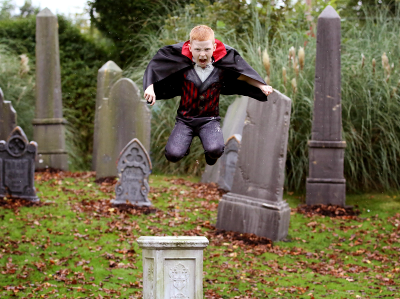 Boy in vampire Halloween costume jumping mid-air among decorative gravestones in an autumn cemetery scene.