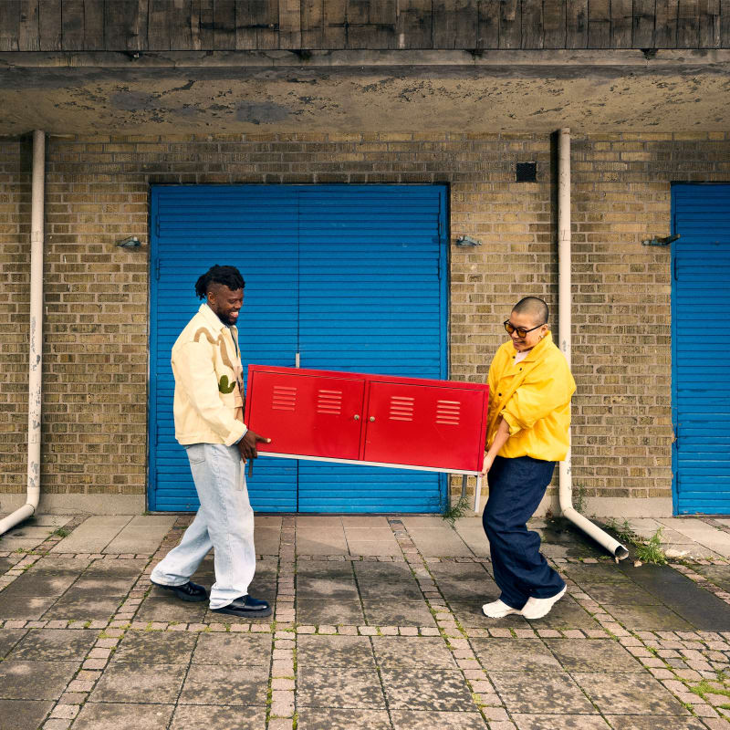 Two people carry a red metal locker cabinet across a cobblestone area in front of blue shuttered doors.