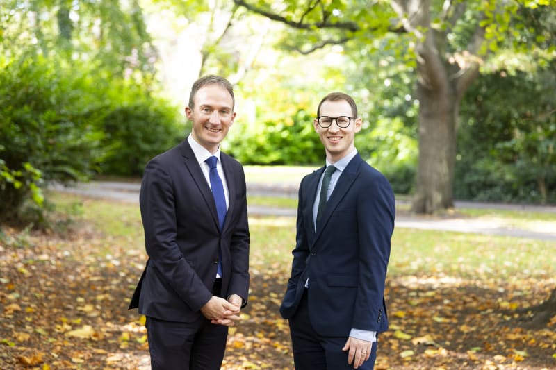 Alan Dillon TD and Adam Isaacs smiling in a park, both wearing dark navy suits.