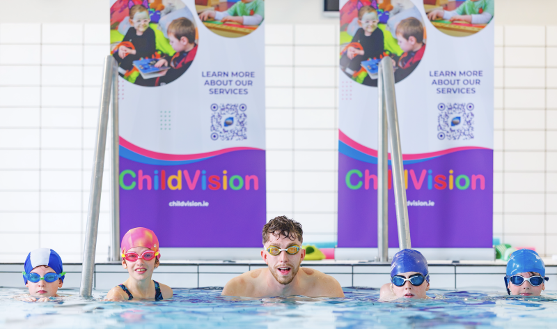 Adult swimmer and four children in goggles and swim caps in an indoor pool in front of ChildVision banners.