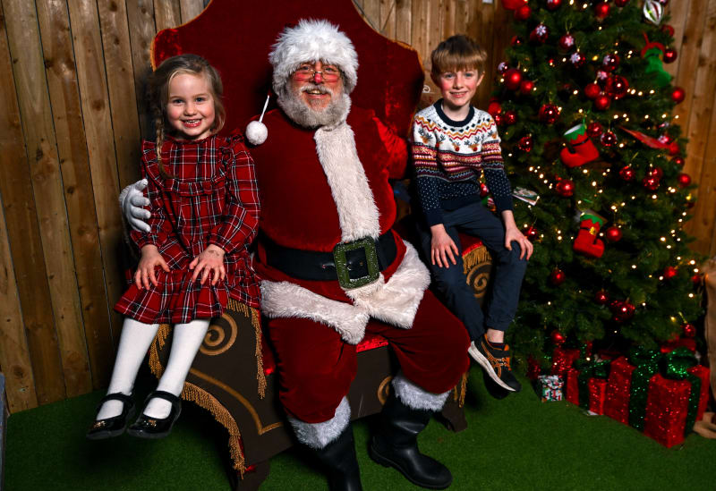 Two children sitting with Santa Claus on a red throne in a Christmas grotto with a decorated tree and gifts.