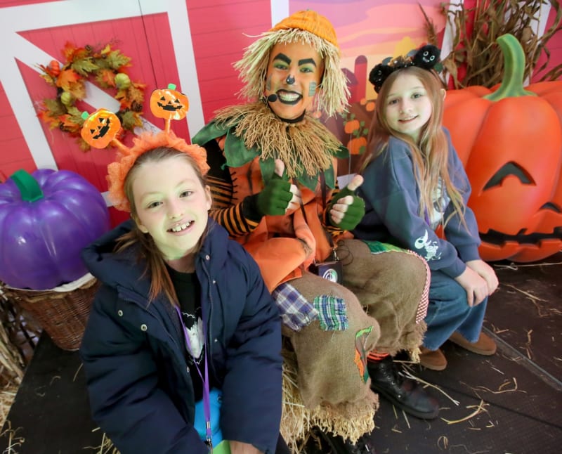 Two girls pose with a scarecrow character performer at a Halloween-themed event with pumpkins and barn backdrop.