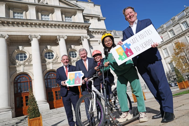Adults and two children with a bike and scooter hold a Safe Routes to School sign outside a civic building.