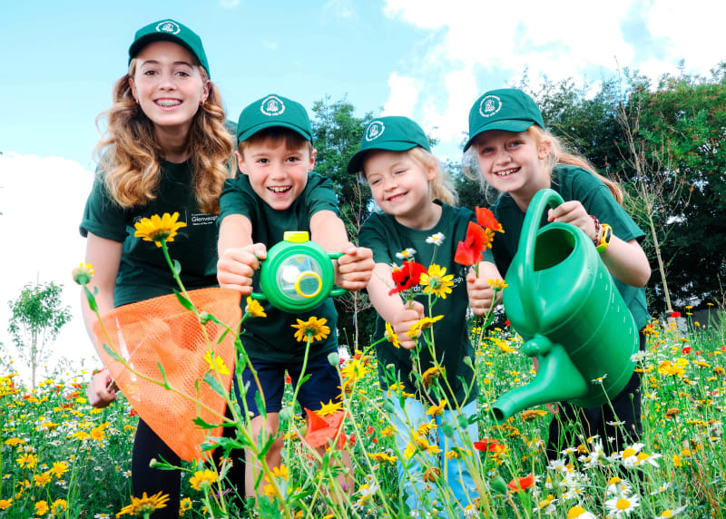 Four children in matching green t-shirts and caps explore a wildflower meadow with a bug viewer, net, flowers and watering can.