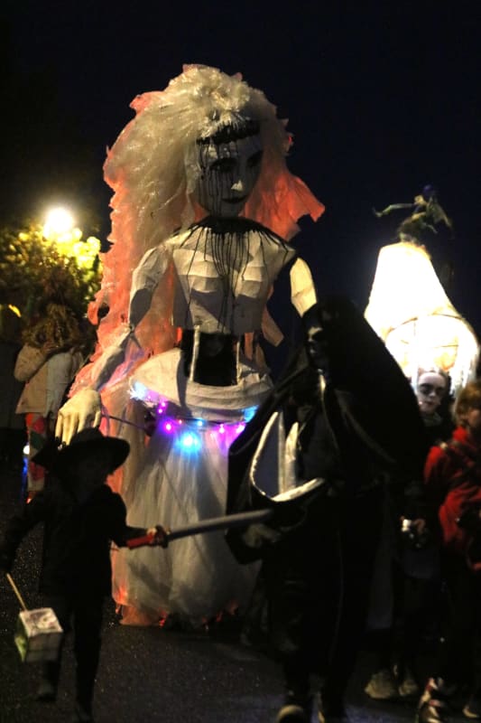 Giant ghostly bride puppet with LED lights at a nighttime Halloween parade, surrounded by costumed participants.