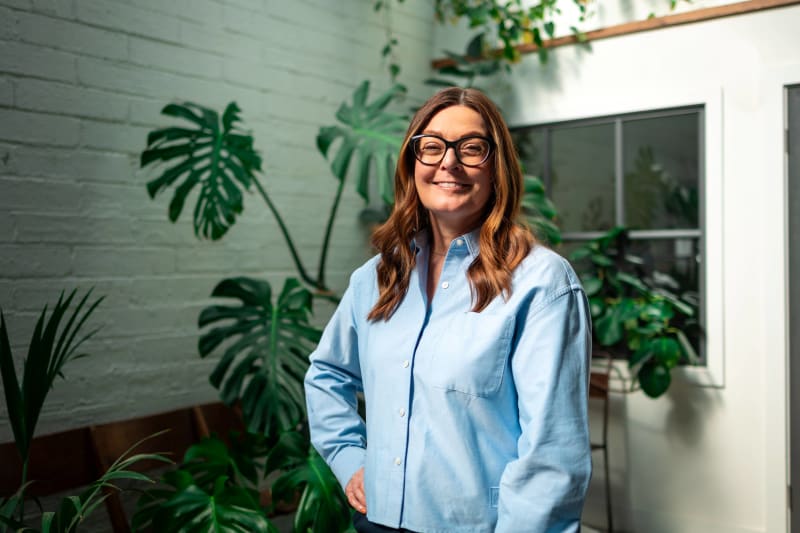Dr Jacqueline De Brún smiling indoors surrounded by large green tropical houseplants.