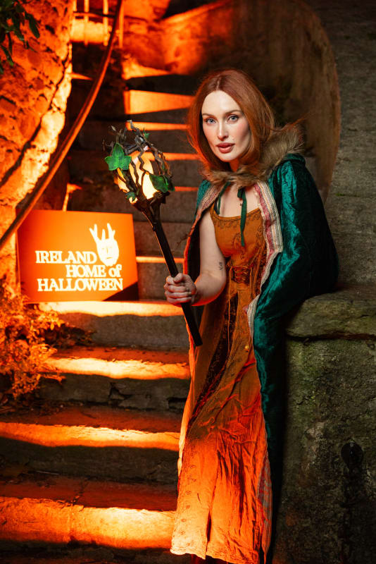 Roz Purcell in Halloween costume holding a glowing staff beside an Ireland Home of Halloween sign.