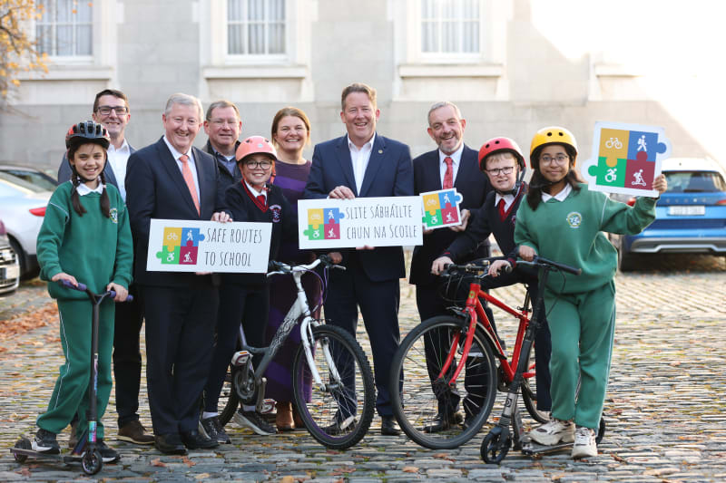 Adults and schoolchildren pose with bikes and scooters holding Safe Routes to School signs at a launch event.