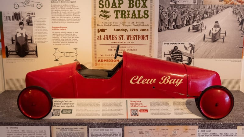 Red vintage soap box derby car named Clew Bay on museum display with 1950s Soap Box Trials poster at Turlough Park.