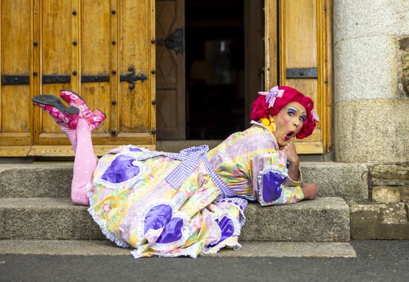 Michael Sands in pantomime dame costume lying on stone steps, wearing pink wig, floral dress and theatrical makeup.